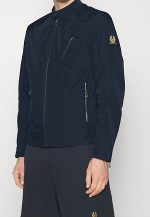 Mand iført navyblå jakke med lynlås, høj krave og lynlåslommer på bryst og sider, kombineret med mørke bukser.