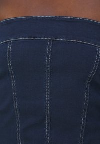 Jeansova bluza v temno modri barvi brez naramnic. Ima belo šivanje in navpične šive za strukturirano obliko. Gladka tekstura.
