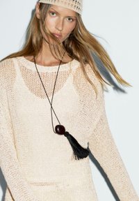 Maglione lavorato a maglia in beige chiaro con trama aperta, dotato di scollatura rotonda e spalle scese. Pendente con nappina nera su una collana nera.