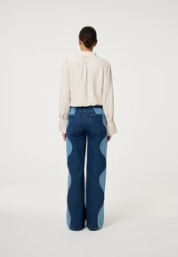 Licht beige blouse met lange mouwen en volume schouders, gecombineerd met hoge taille spijkerbroeken. De spijkerbroeken hebben golvende lichtblauwe denim accenten.