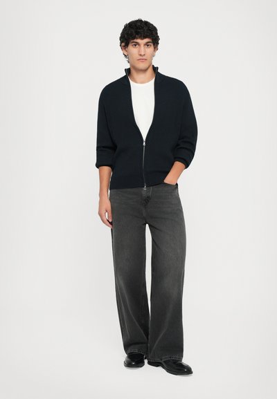 Marinblå ribbad zip-up tröja med korta �ärmar, i kombination med lösa svarta jeans och svarta skor, mot en vit bakgrund.