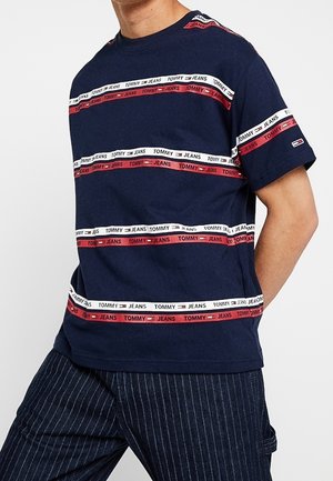 Man wearing navy blue Tommy Jeans t-shirt with red and white horizontal logo stripes and dark pinstriped pants, standing with hands behind back.
