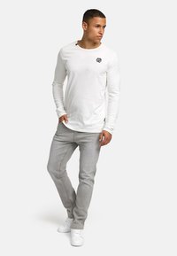 Chemise blanche à manches longues avec un col rond et un petit logo noir ; associée à un jean slim gris clair et des baskets blanches.