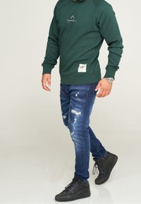 Uomo che indossa una felpa verde scuro e jeans blu strappati, sneakers nere alte e un orologio da polso su uno sfondo semplice.
