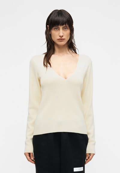 Pull crème à col en V avec manches longues, texture douce et motif tricot subtil. Accompagné de pantalons larges noirs et d'un détail d'étiquette blanche.
