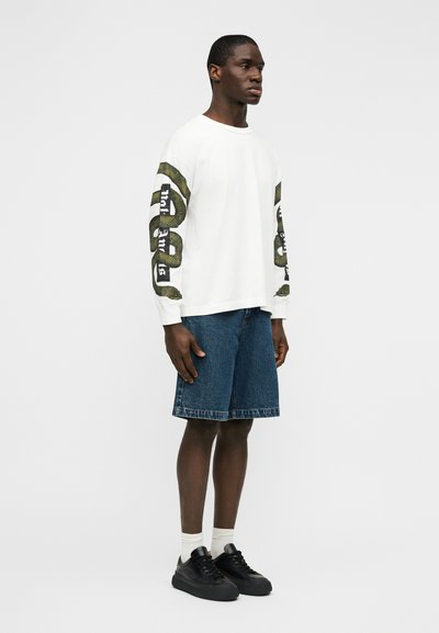 Μπλούζα με μακριά μανίκια σε λευκό χρώμα με πράσινο γραφικό φίδι στα μανίκια, συνδυασμένη με μπλε τζιν σορτς. Φορώντας μαύρα αθλητικά παπούτσια και λευκές κάλτσες.