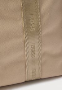 Beżowa torba z tkaniny o fakturowanej powierzchni, plecionym pasku z wytłoczonym logo oraz gładkim wykończeniu.