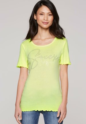 Woman wearing a bright yellow short-sleeve top with scalloped edges and text design, paired with blue jeans, standing against a plain background.