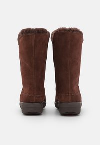 Bottes en daim marron avec une doublure en fourrure douce au sommet, présentant un bout arrondi et une semelle en caoutchouc, avec des coutures visibles sur les bords.