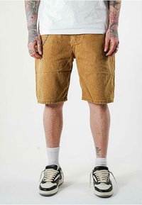 Pantaloni corti di velluto a coste color corda con tasche laterali, caratterizzati da un taglio dritto e cuciture in vista. Abbinati a sneaker nere e bianche e calzini bianchi.