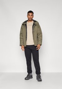 Olivgrüne isolierte Jacke mit Kapuze, beiges T-Shirt, schwarze Jogginghosen und graue Sneaker, alles in einer frontalen Pose vor einem weißen Hintergrund.