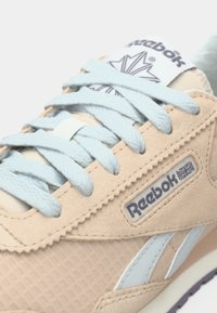 Tuvošs plāns skats uz bežām Reebok sporta apavām ar gaiši ziliem šņorēm un logo tagu, uz kura redzama Lielbritānijas karoga attēls uz teksturētas auduma.