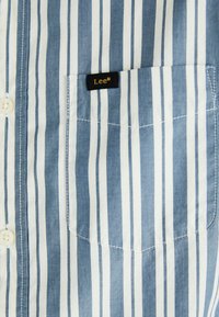 Chemise à rayures bleues et blanches avec des boutons et une poche poitrine. Présente une étiquette de marque Lee® en noir et or sur la poche. Tissu en coton lisse.
