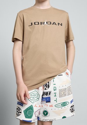Personne portant un t-shirt Jordan beige et un short blanc avec différents logos graphiques colorés et des motifs de texte.