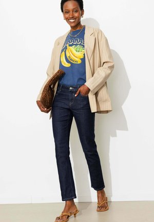 Femme souriante portant un blazer beige oversize, un t-shirt bleu avec un motif banane, un jean foncé, des sandales sable et tenant une pochette marron à motif.