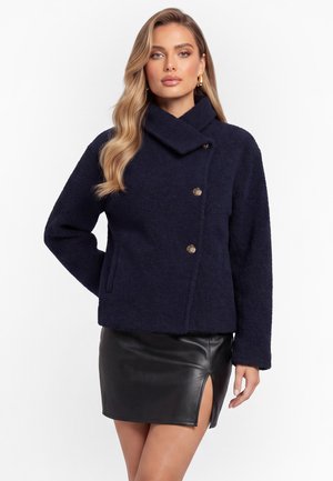 Frau mit langen, welligen blonden Haaren, die eine marineblaue, strukturierte Jacke mit goldenen Knöpfen und einen schwarzen Leder-Minirock mit einem Frontschlitz trägt.