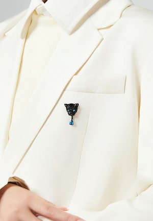 Spilla a forma di pantera nera con accenti blu, caratterizzata da dettagli intricati e un cristallo a goccia, appuntata a una tasca di un blazer bianco con texture.