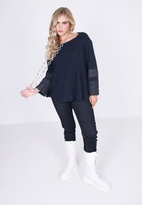 Marineblauwe chunky gebreide trui met kanten accenten op de mouwen, gecombineerd met donkere denim broek en witte knielange veterlaarzen met een zilveren ketting.