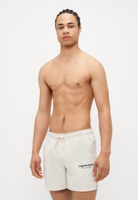 Beige badeshorts med elastisk midje, justerbar snøring og lomme på venstre side. Har sort logo-trykk foran.