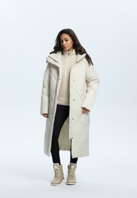 Langer, elfenbeinfarbener Puffermantel mit einem gesteppten Muster, großer Kapuze und Druckknöpfen. Kombiniert mit einem beigen Pullover, schwarzen Hosen und hellbraunen Stiefeln.