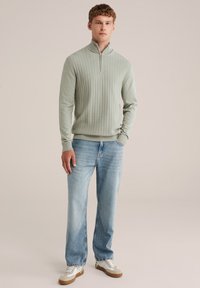 Gaiši zaļa ribota džemperis ar rāvējslēdzēja kaklu, kombinēts ar gaiši zilām taisnākājām džinsām un baltām sporta apaviem ar brūniem akcentiem. Vienkāršs, teksturēts dizains.