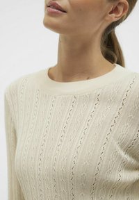 Maglione lavorato a maglia di un beige chiaro con un motivo a treccia rigato e piccoli fori. Il collo presenta una forma morbida e arrotondata.