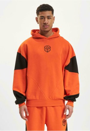 Oranž kapuutsiga sü hoodie, millel on mustad aktsendid, esiosas logo ja soonikkoes. Valmistatud pehmest kangast, demonstreerides mugavat lõiget ja geomeetrilisi õmblusi.