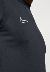 Haut noir ajusté en tissu lisse avec un logo de marque blanc brodé sur la poitrine. Design simple sans motifs supplémentaires.