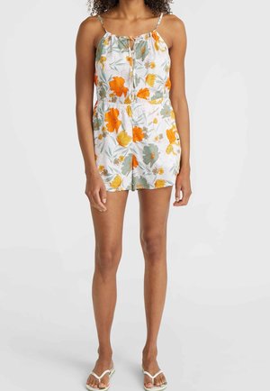 Woman wearing a sleeveless romper with orange and yellow floral print, spaghetti straps, front tie detail, and pockets, paired with white sandals.