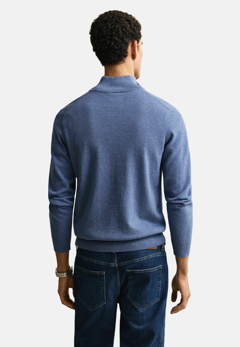 Mann trägt einen blauen Pullover mit langen Ärmeln und dunkelblaue Jeans, steht mit dem Rücken zur Kamera und trägt eine Uhr am linken Handgelenk.