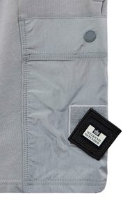 Grått stoff med en lomme og en rund knapp. En svart label med teksten "WEEKEND OFFENDER" er delvis synlig, med en myk teksturbakgrunn.