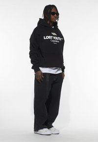 Felpa nera con la scritta "LOST YOUTH", maglietta bianca sotto, pantaloni in denim scuro e sneakers bianche con suole grigie. Sono visibili occhiali da sole e treccine.