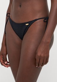 Black bikini bottom with ribbed texture, low rise, adjustable ties, and a small gold logo accent on the side.