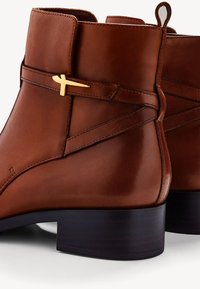 Bottines en cuir marron avec une finition lisse, une fine sangle latérale avec des détails en doré et un petit talon bloc.