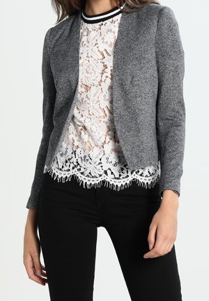 Frau trägt eine graue offen vorn offenen Strickjacke über einem weißen Spitzenoberteil mit floralen Mustern, kombiniert mit schwarzen Hosen.