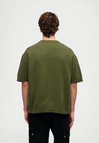 Μπλούζα από πλεκτό, με κοντά μανίκια σε ελαιόλαδο χρώμα, με στρογγυλή λαιμόκοψη, που διαθέτει ριγωτές μανσέτες και τελείωμα, εμφανιζόμενη από πίσω.