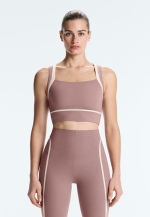Kvinde stående med fronten mod kamera iført mauve sports-BH og høj talje leggings med beige kant, mod ensartet lyst baggrund.