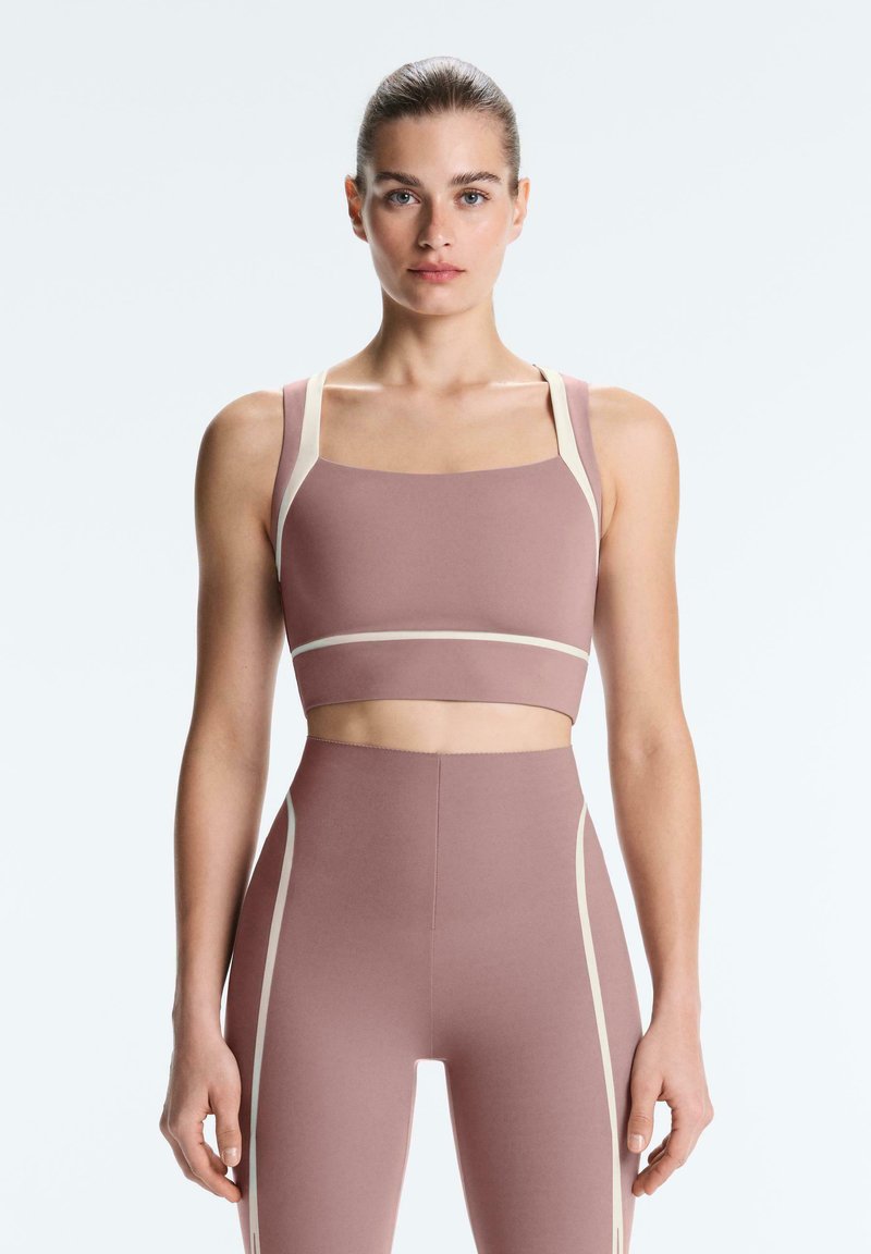 Femme debout face à l’objectif, portant un brassard de sport mauve et des leggings taille haute avec un bord beige, sur un fond clair uni.