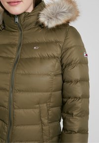 Olivgrön pufferjacka med dragkedja, elastiska muddar och avtagbar huva med pälsfoder. Har en logotyppatch på vänster arm.