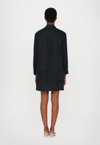 Black long-sleeve shirt with a back pleat, paired with a matching skirt. Material appears textured. High-heeled shoes in a nude tone.