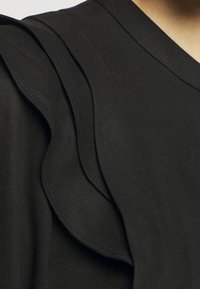 Haut noir en tissu texturé avec des détails à volants superposés le long de l'épaule et du col, créant un intérêt visuel.