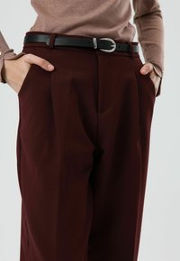 Bordeauxkleurige, op maat gemaakte broek met een gladde textuur, voorzien van plooien aan de voorkant en een zwarte lederen riem met een zilveren gesp.