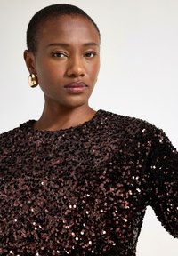 Sort pailletetop med rund halsudskæring, der har en tekstureret overflade og subtil glans. Guldøreringe i hoops fremhæver looket.