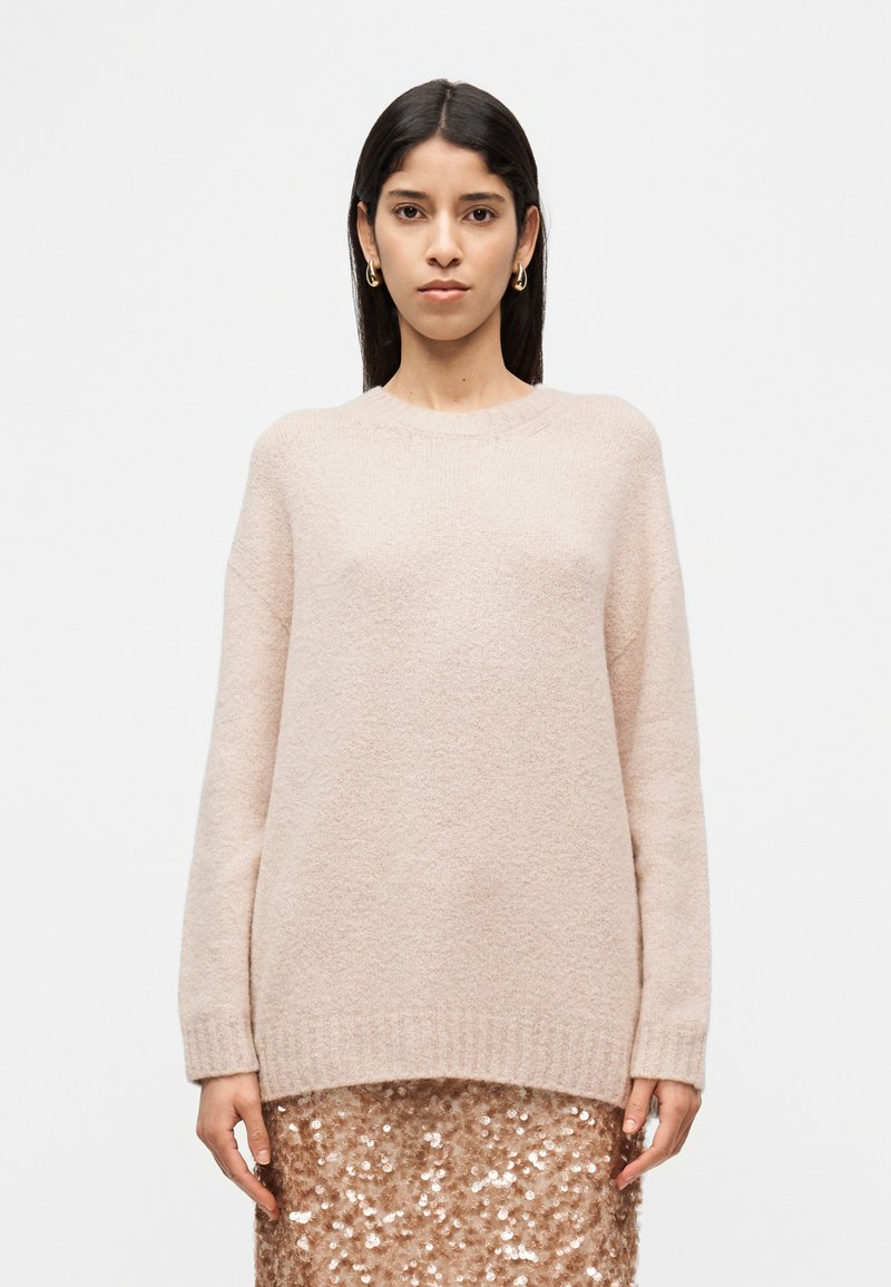 Παντελόνι σε ροζ ανοιχτό χρώμα, φτιαγμένο από υλικό πλεκτό με υφή, με στρογγυλή λαιμόκοψη και ριμπ μανσέτες, φορεμένο πάνω από φούστα με παγιέτες.