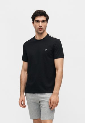 Mies, joka pukeutunut mustaan lyhythihaiseen T-paitaan, jossa pieni logo rinnassa, ja harmaisiin shortseihin, seisoo yksivärisen valkoisen taustan edessä.