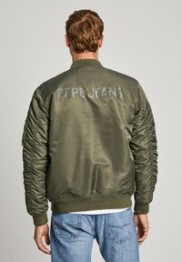 Chaqueta bomber verde olivo con mangas acolchadas y dobladillo acanalado, con la inscripción "PEPE JEANS" en letras mayúsculas de color gris claro en la parte posterior.