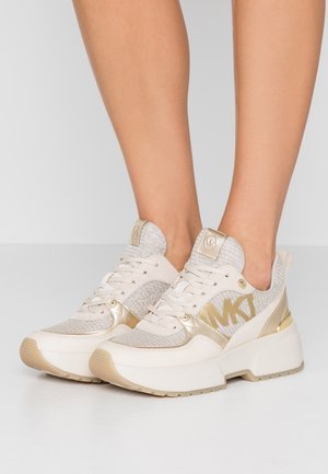 Beige og guld platformsneakers med mesh og læderdetaljer, båret på bare fødder, der viser side-MK-logo og tykke såler.