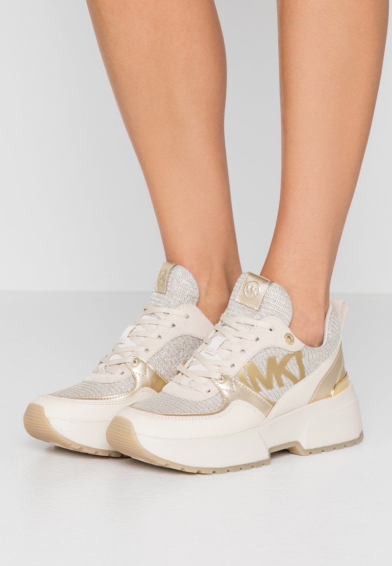 Baskets à plateforme beige et or avec détails en mesh et cuir, portées pieds nus, affichant le logo MK sur le côté et des semelles épaisses.