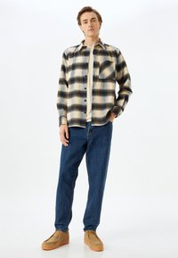 Skjorte i ternet flannel i creme, sort og gul med en brystlomme, parret med blå jeans og brune sko. Glat stofstruktur.