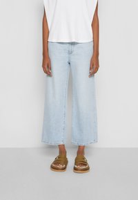 Jeans de mezclilla de pierna ancha en azul claro, combinados con una blusa blanca de canalé y sandalias marrones con tiras y suela gruesa.